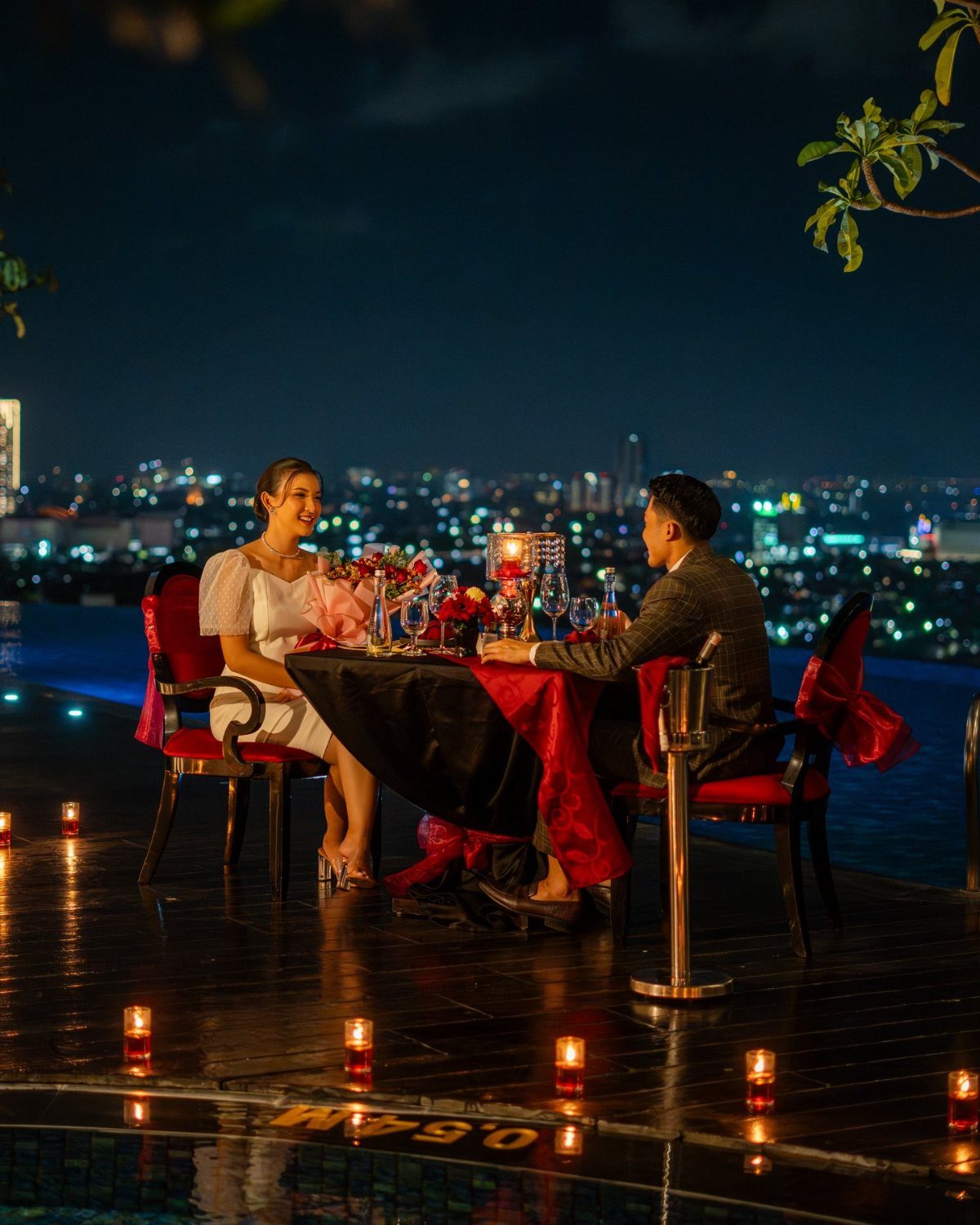A couple enjoys a romantic candlelit dinner on a poolside deck overlooking a city skyline.