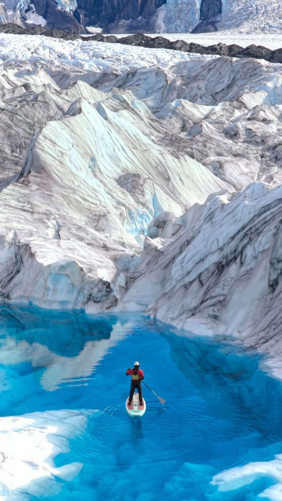 A person paddleboarding on vibrant blue glacial water surrounded by massive ice formations.