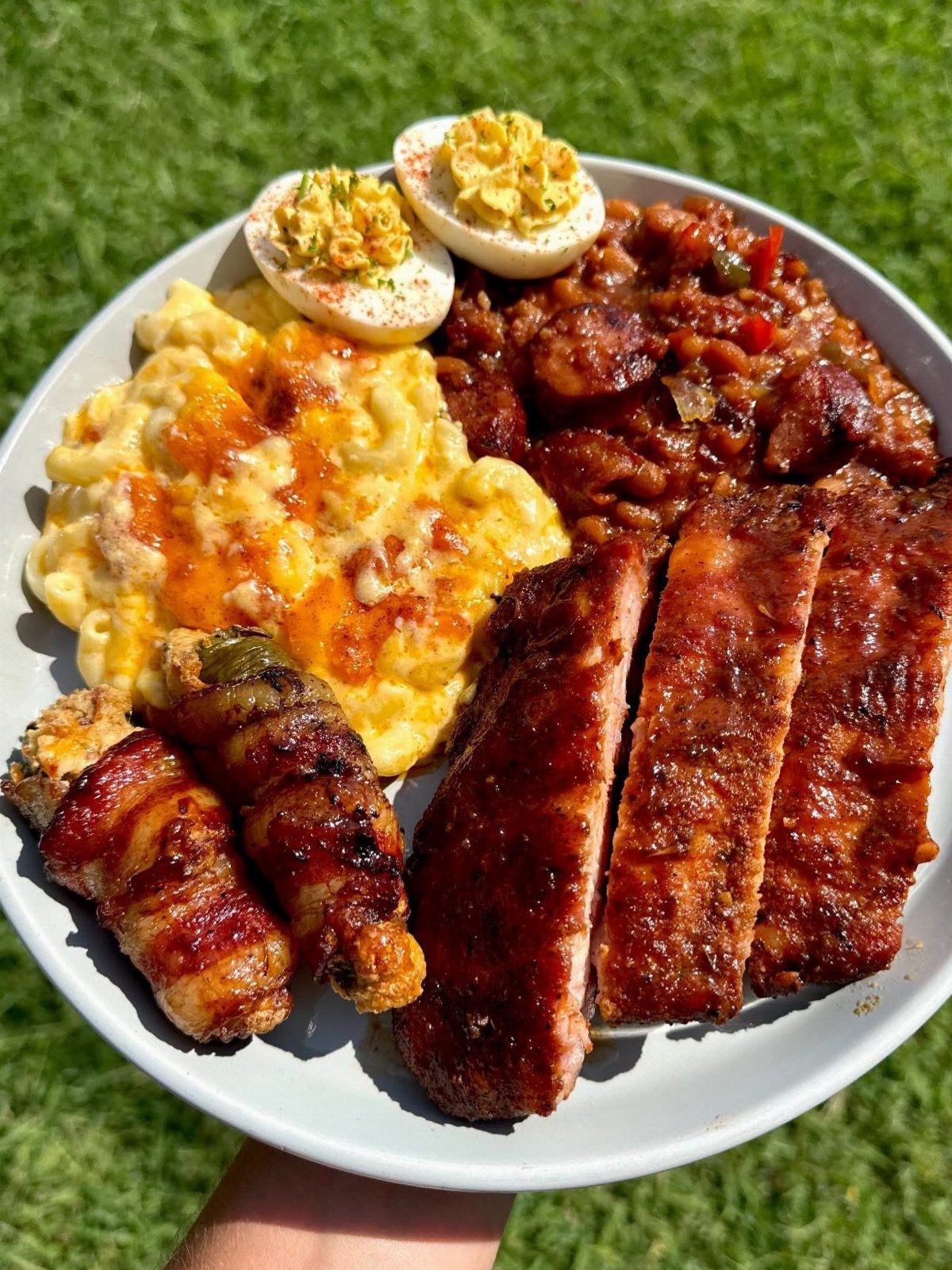 A plate of gourmet barbecue food featuring ribs, mac and cheese, and deviled eggs held over grass.