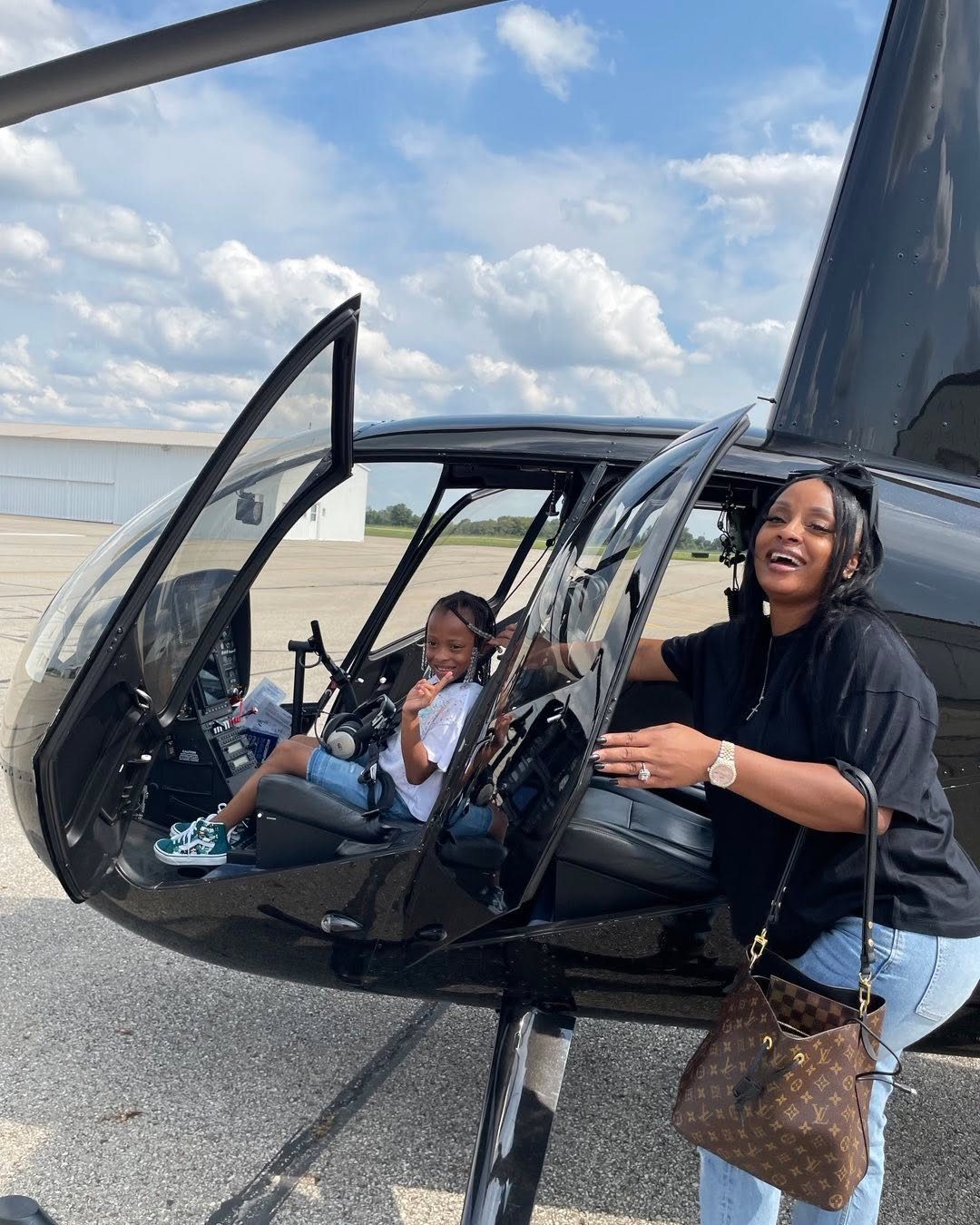 A woman smiling next to a black helicopter with a child sitting inside the cockpit.