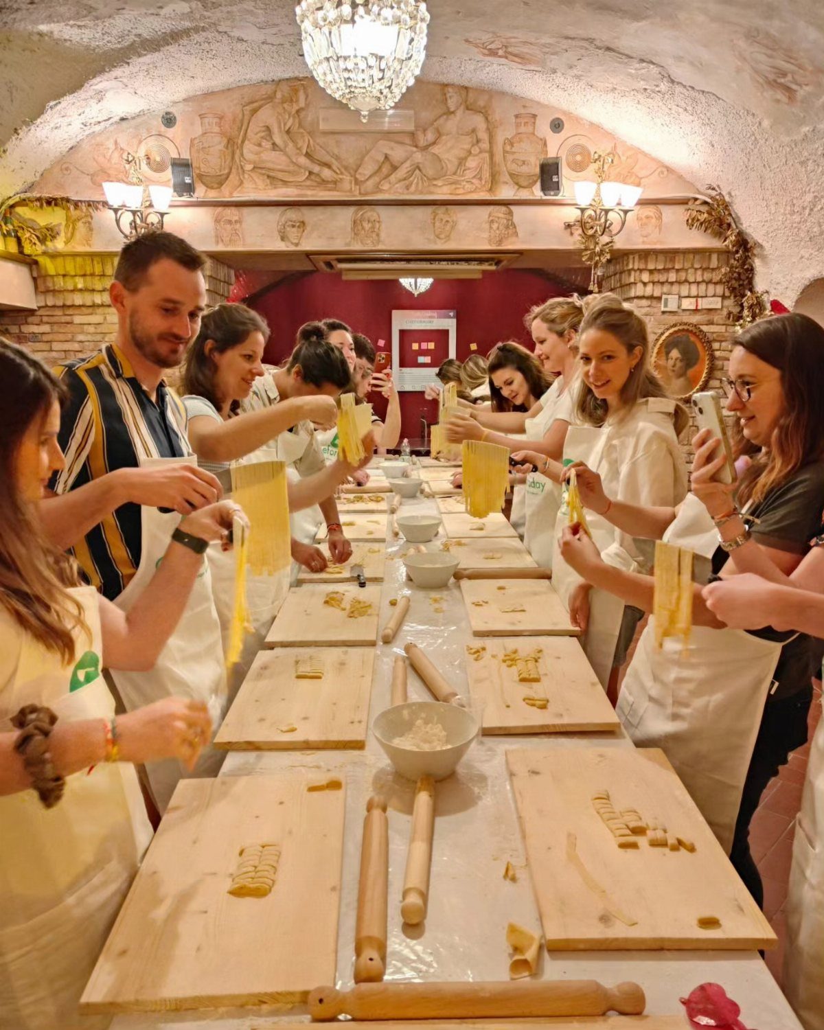 People in white aprons making fresh pasta together at a long wooden table.