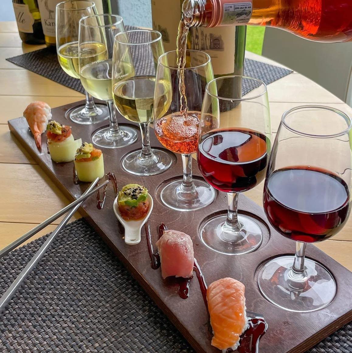 Wine glasses and sushi pieces arranged on a wooden serving platter outdoors.