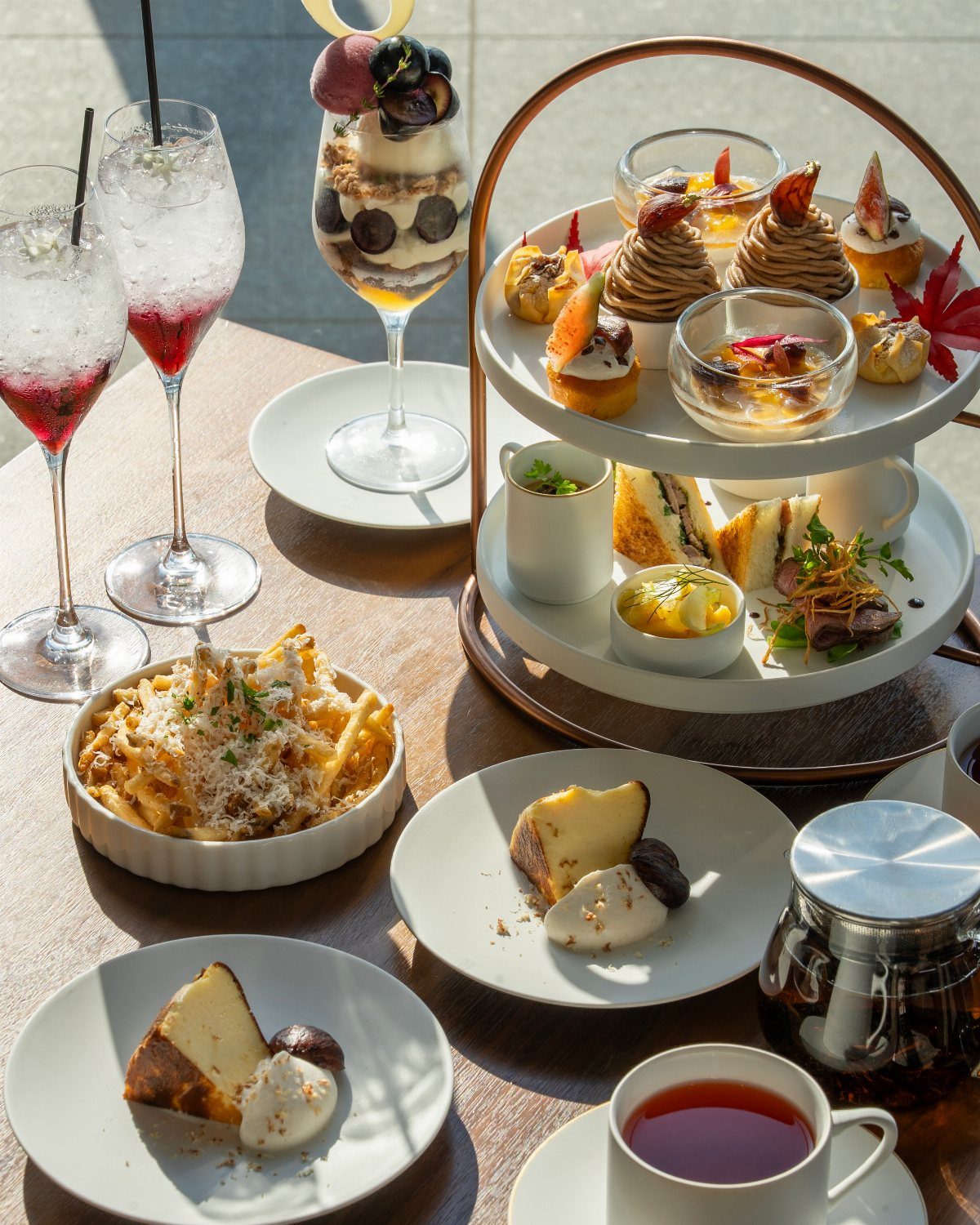 A tiered afternoon tea stand with pastries and sandwiches next to fruit parfaits and tea.