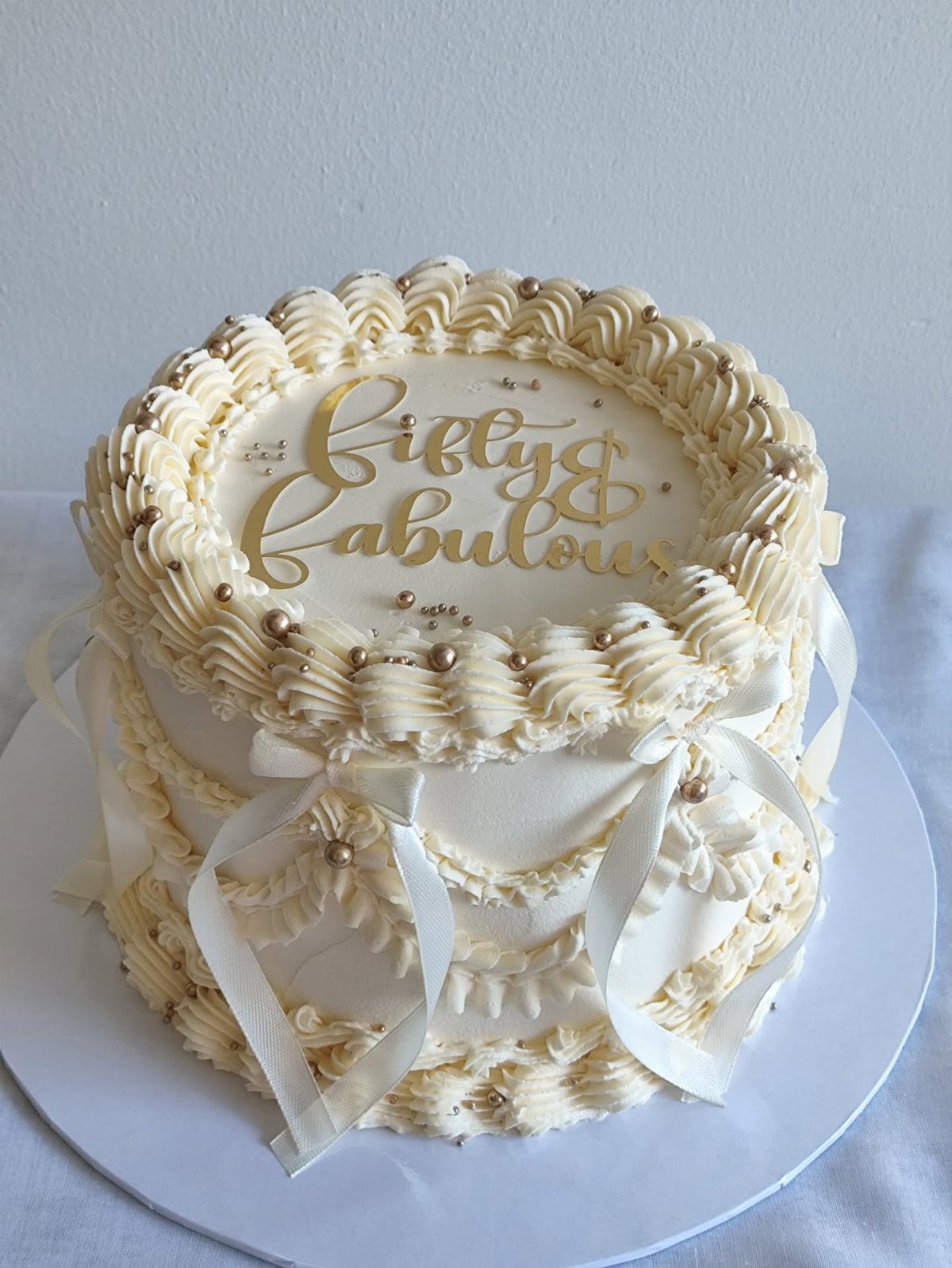 A white frosted cake with gold lettering reading Fifty and Fabulous and decorative ribbons.