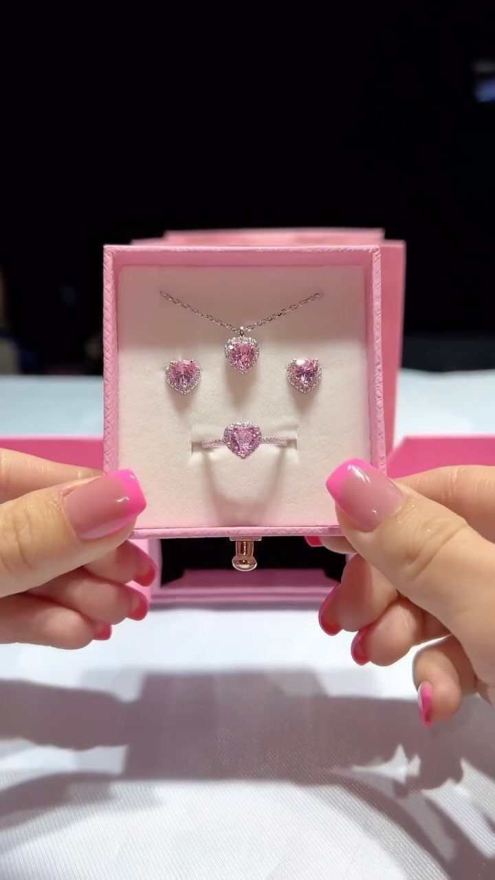 Hands holding an open pink jewelry box containing a heart-shaped silver and pink gemstone set.