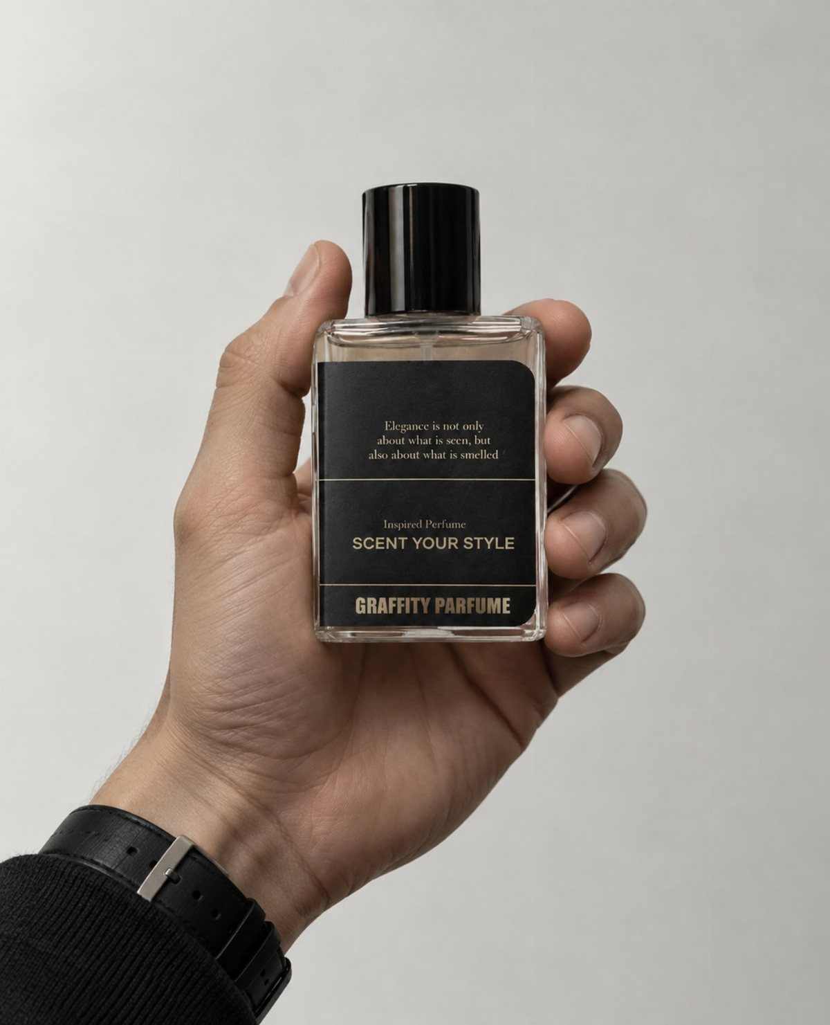 A hand holding a clear glass perfume bottle with a black label against a grey background.