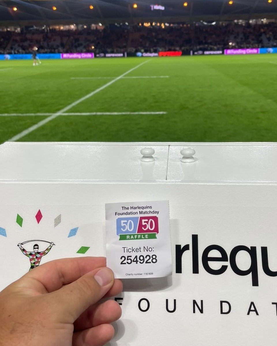 A hand holding a white 50/50 raffle ticket in front of a rugby stadium.