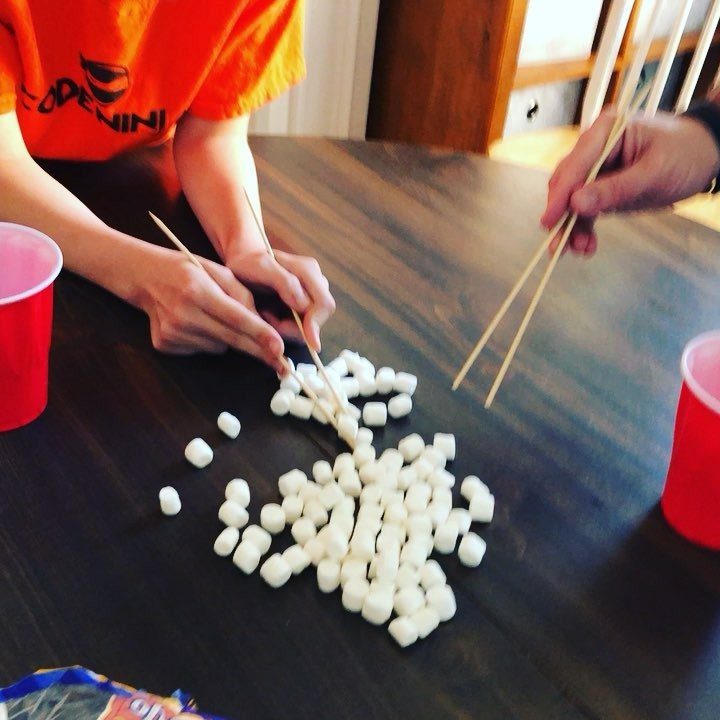 Hands using wooden skewers to move white marshmallows on a dark wooden table.