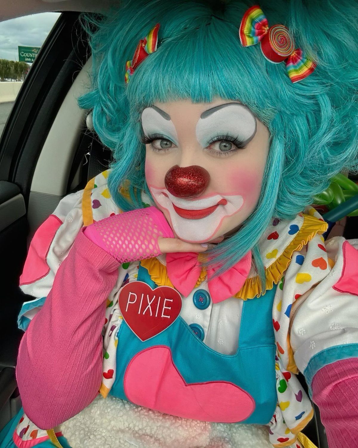 A person in colorful clown makeup and a teal wig posing in a car.