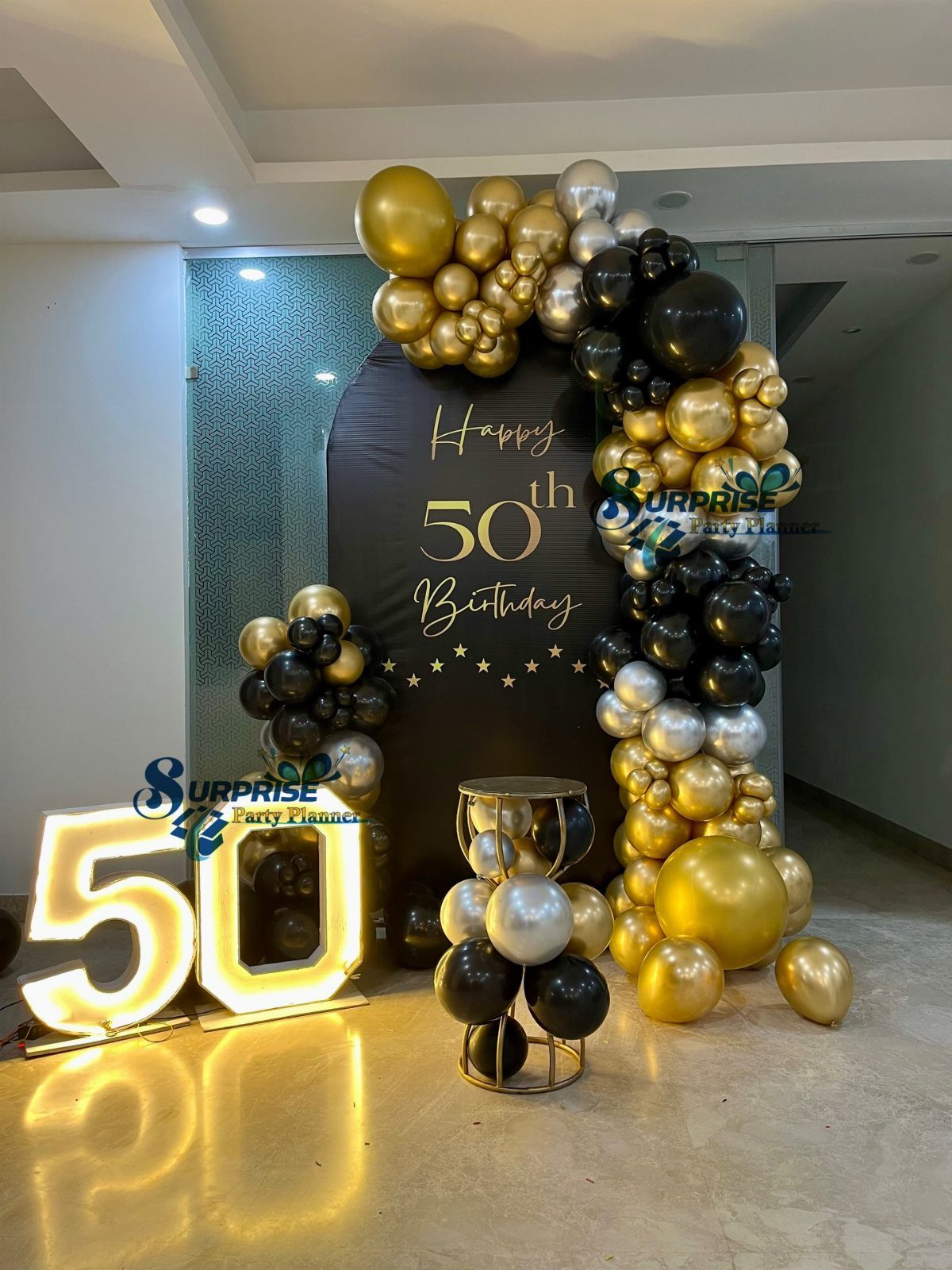Gold, black, and silver balloon arch surrounding a black 50th birthday backdrop and lighted numbers.