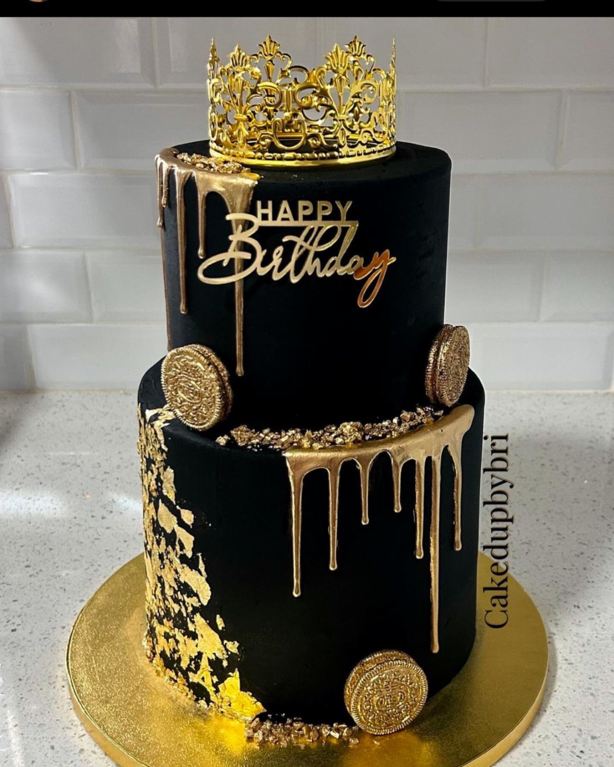 Two-tier black cake with gold drips, gold macarons, and a gold crown topper.