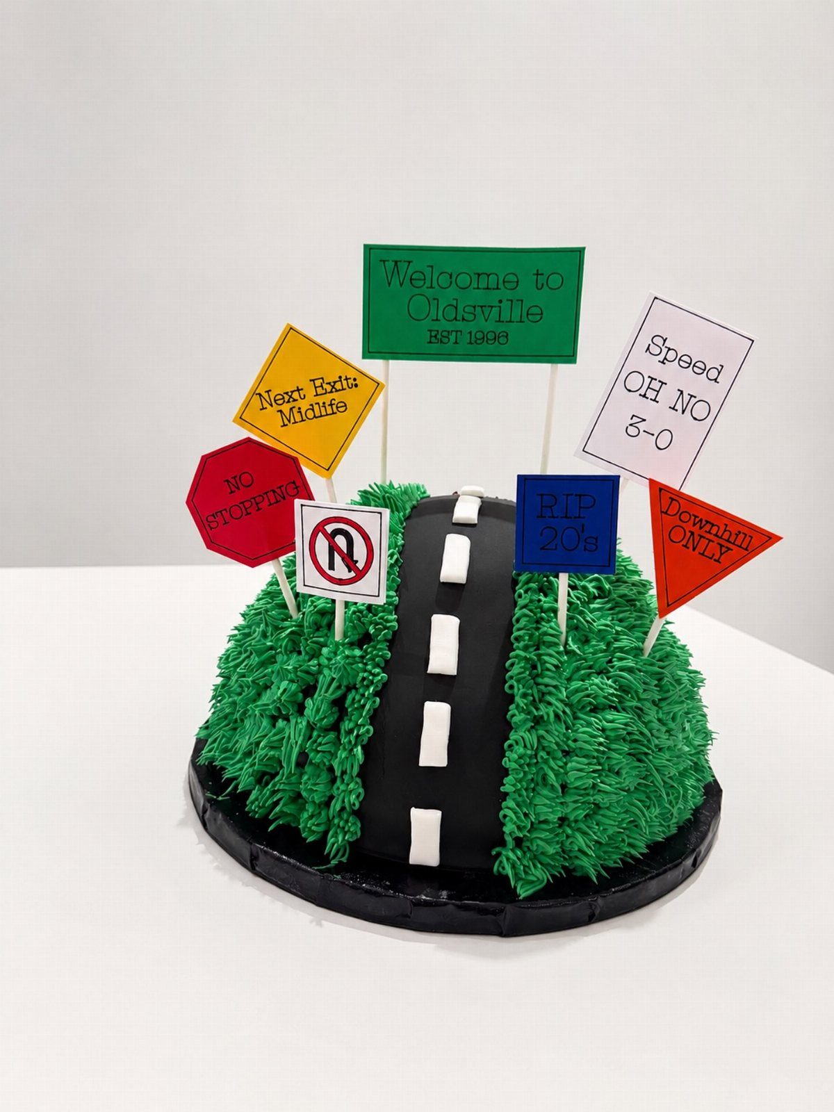 A novelty cake featuring a winding road and humorous road signs on a green grassy hill.