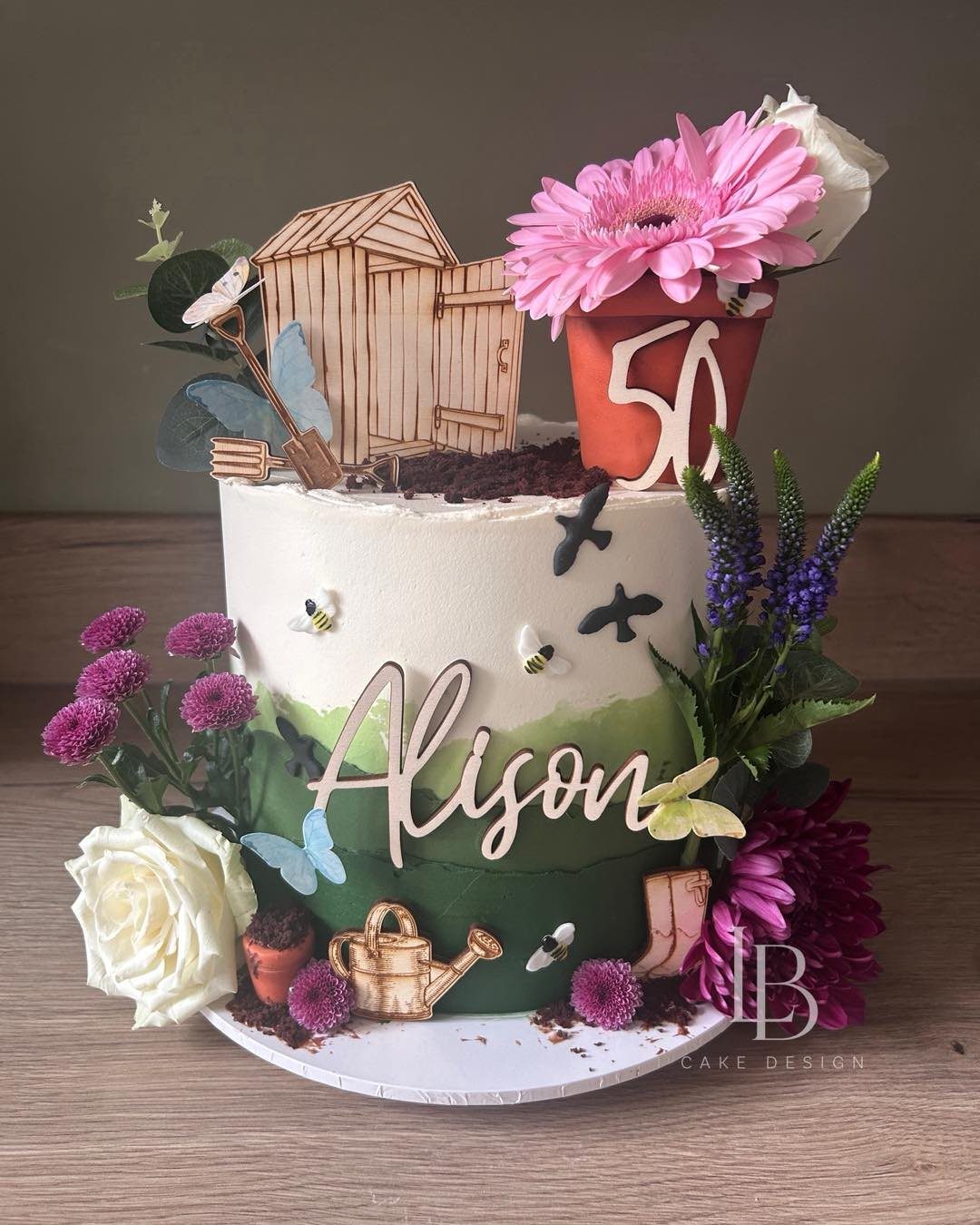 A garden themed birthday cake decorated with flowers, a wooden shed, and a watering can.