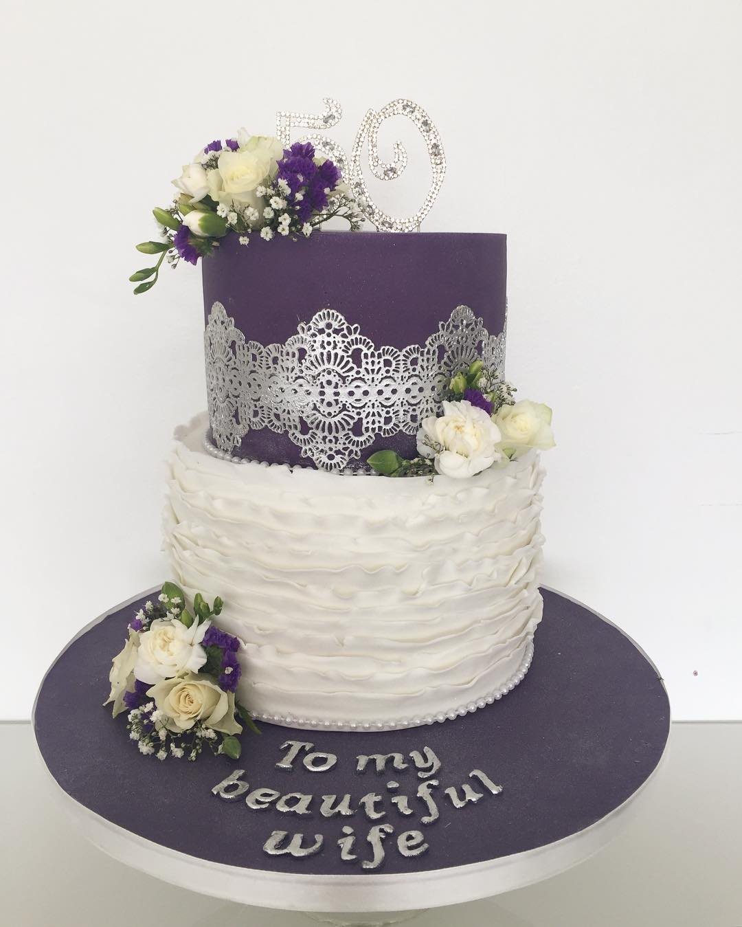 Two-tier purple and white cake decorated with silver lace, flowers, and a fifty topper.