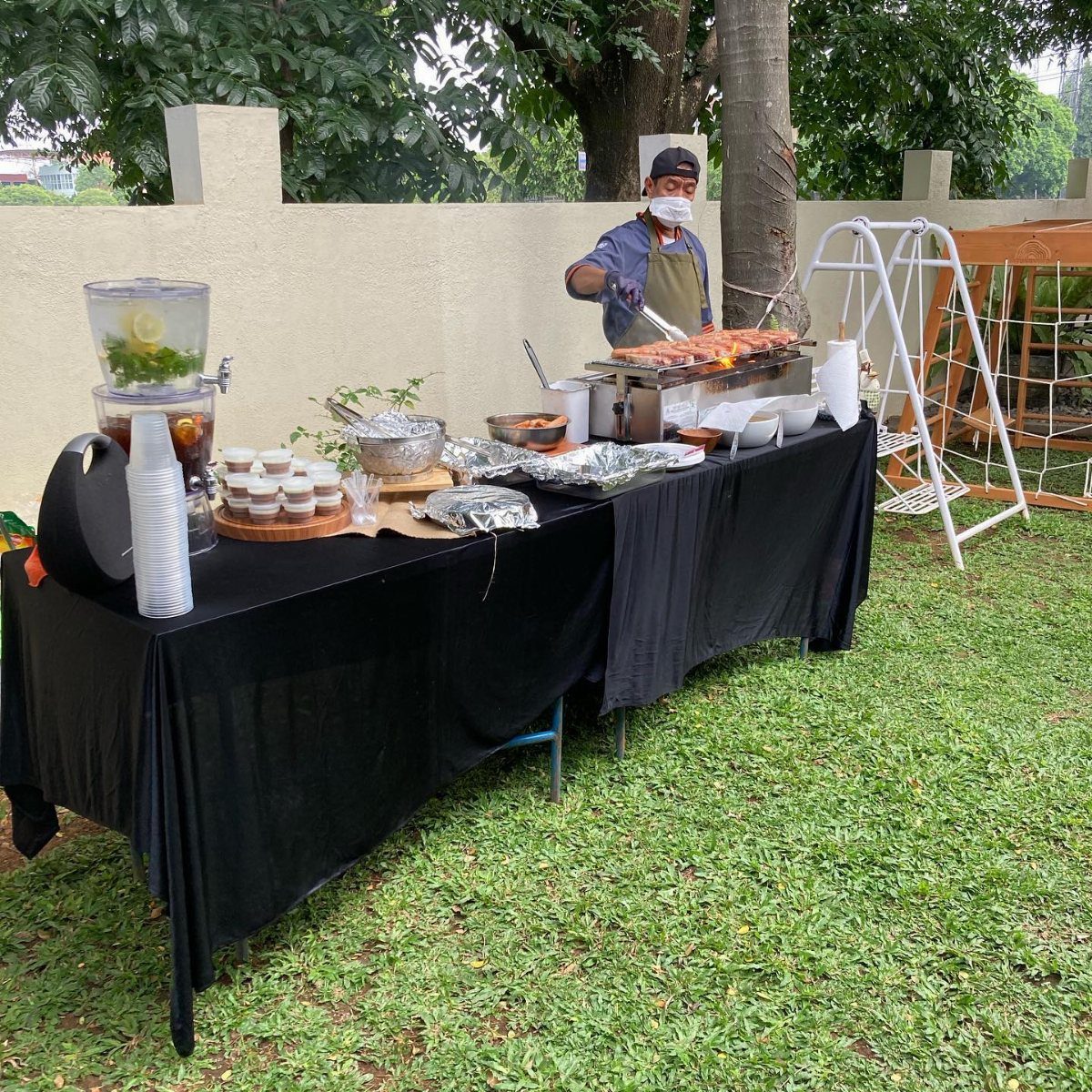 A man grilling food at an outdoor buffet table set on a green lawn.