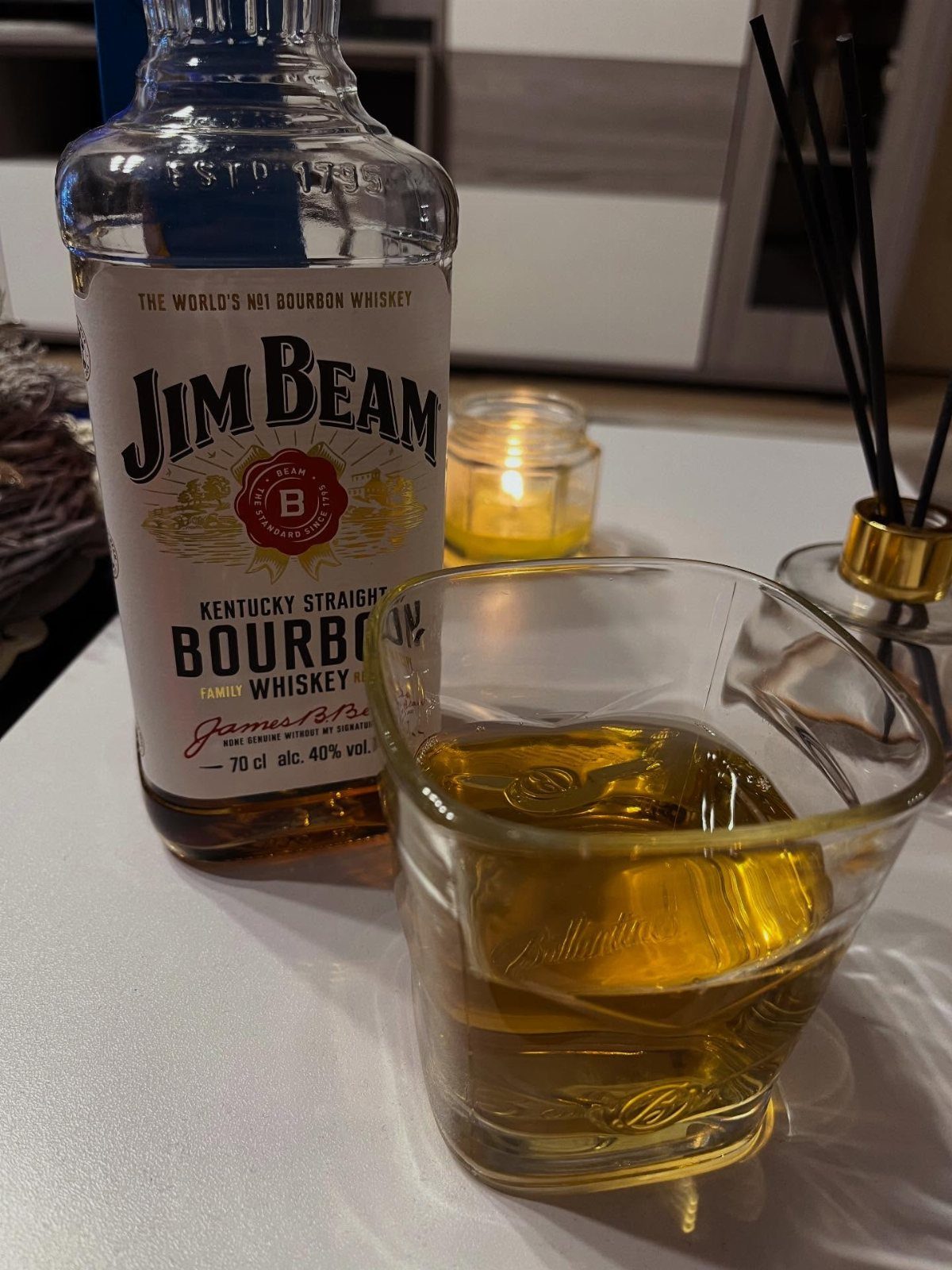 Jim Beam bourbon bottle and a glass of whiskey on a white table with a candle.