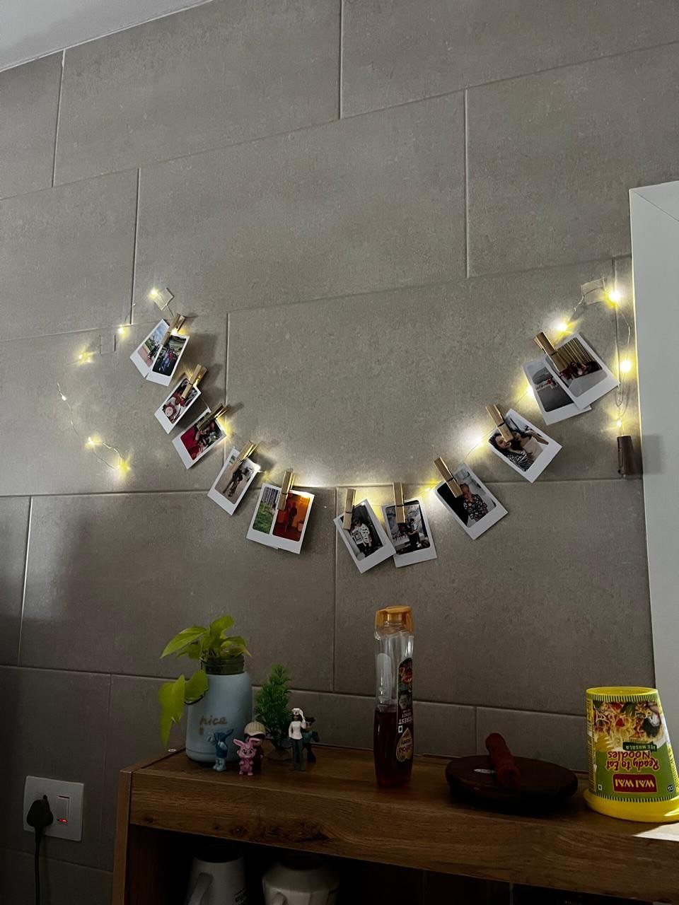 Instant photos clipped to a string of glowing fairy lights on a grey tiled wall.