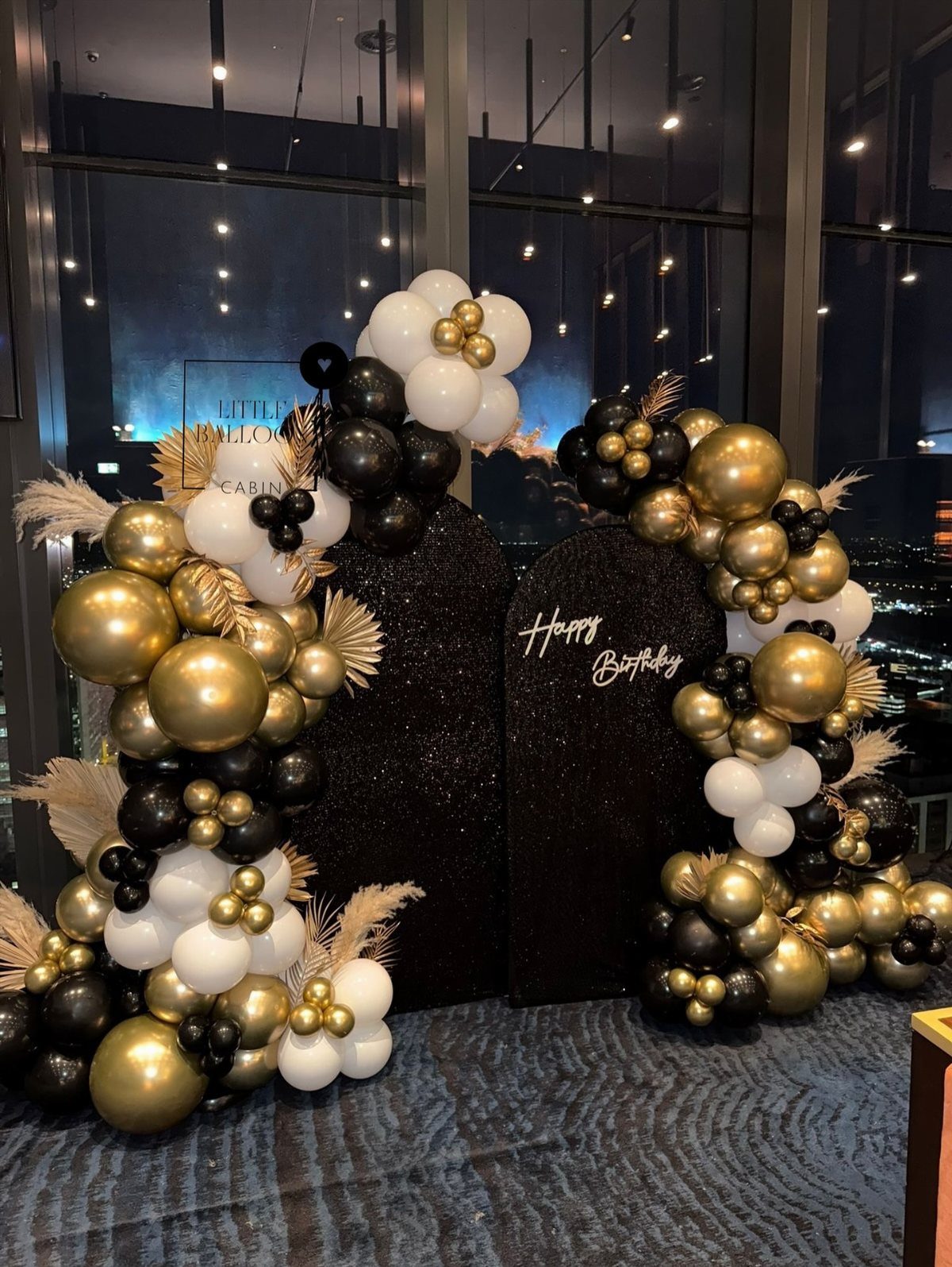 Black glittery arch backdrop decorated with gold, black, and white balloon clusters against a city skyline.