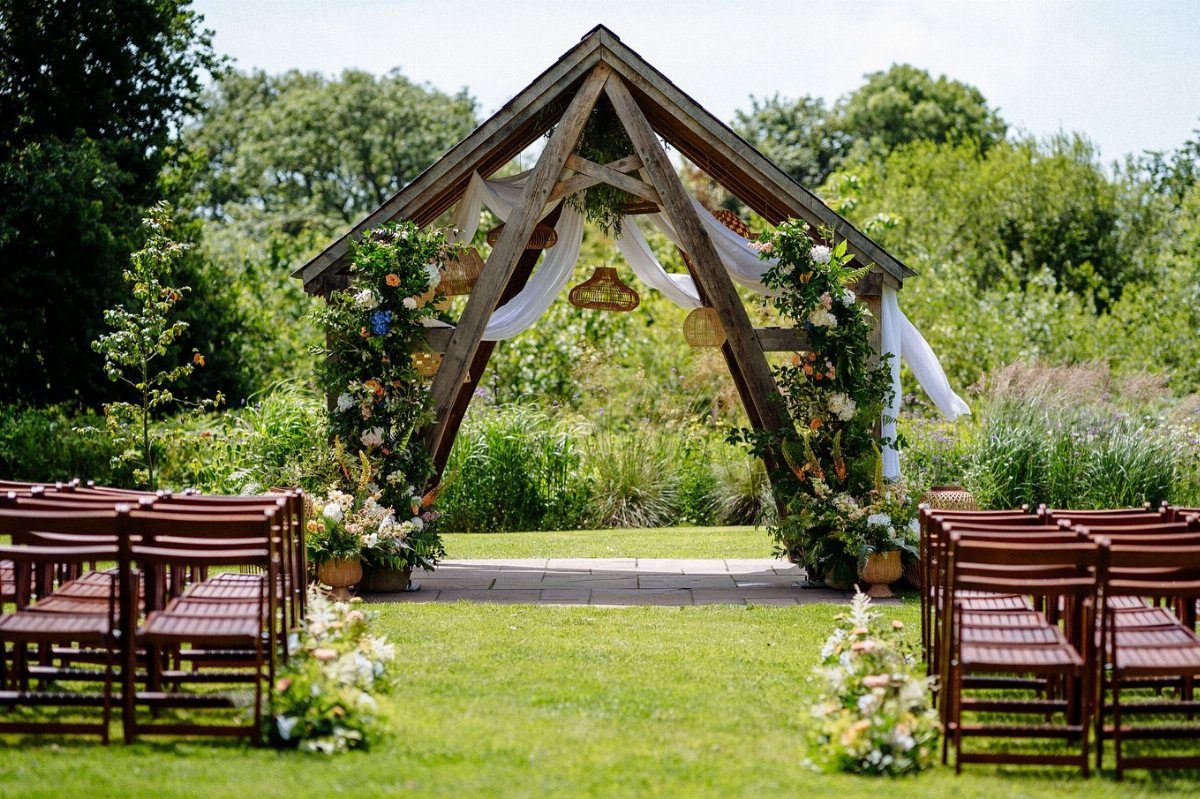 Wooden A-frame arch decorated with white fabric and flowers in a lush green garden.