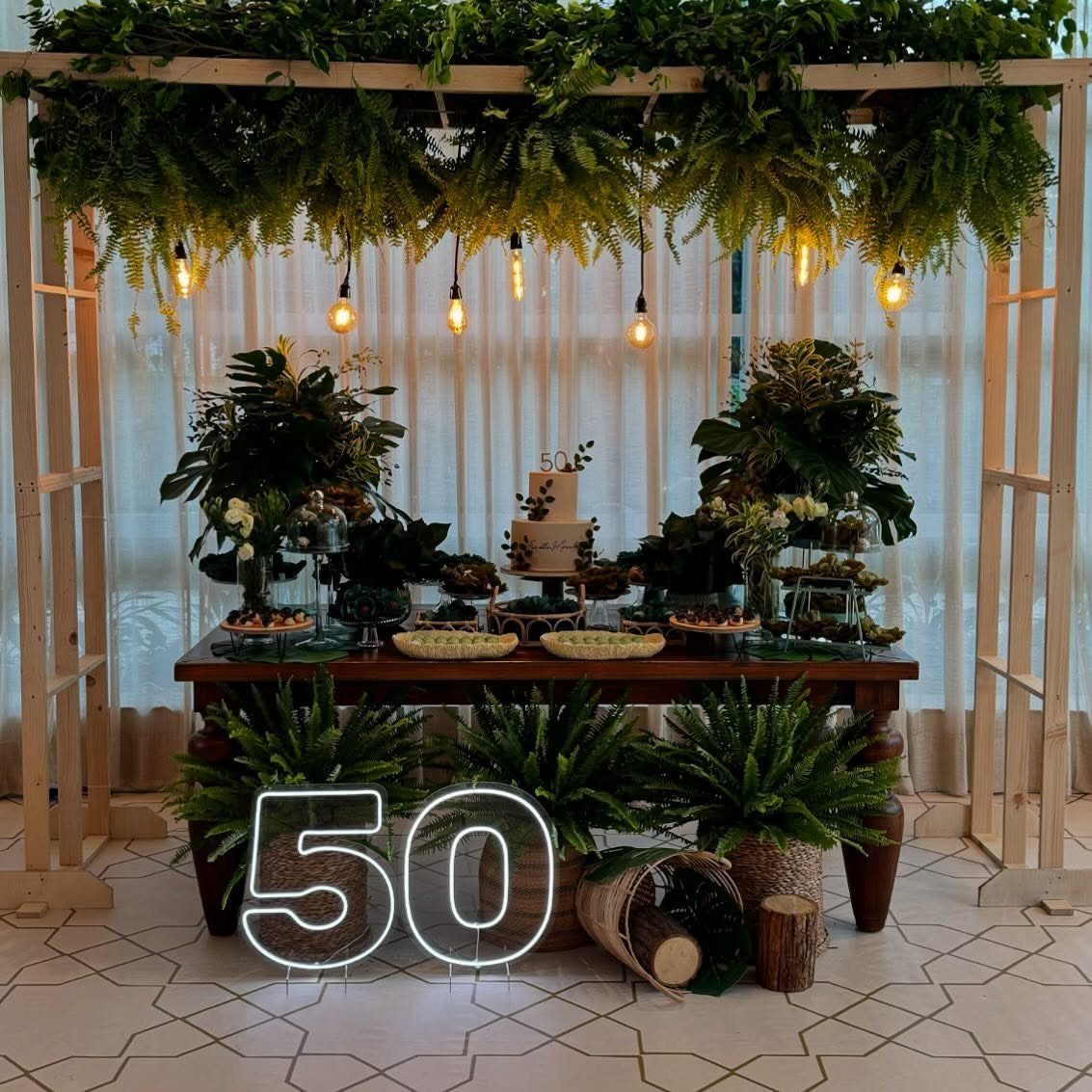 A decorated buffet table with a white cake, greenery, and a large neon number 50 sign.