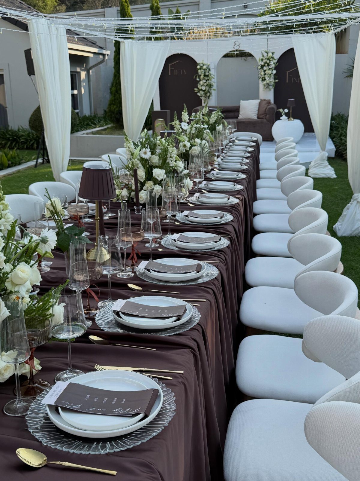 A long formal dining table set with white plates and flowers under a white canopy.