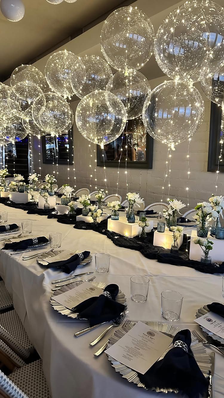 A long banquet table decorated with white flowers, black napkins, and floating light-filled balloons.