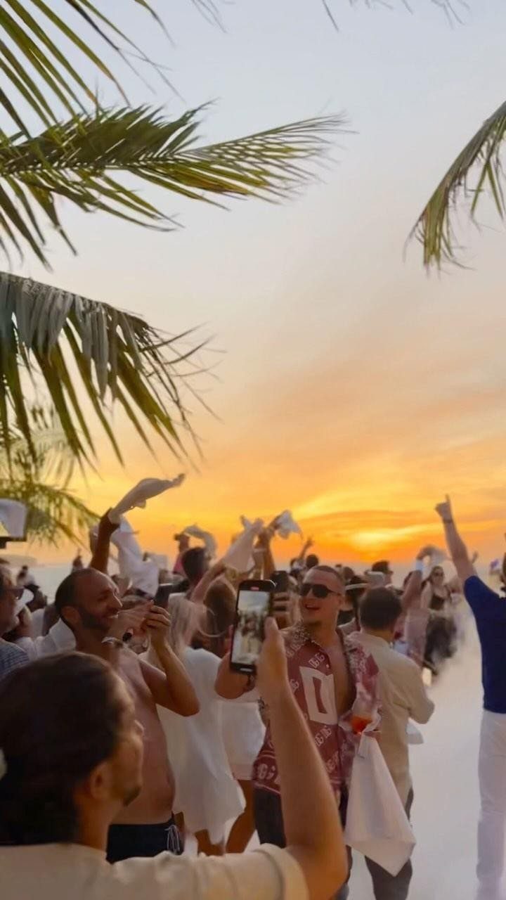 People in white outfits celebrating on a sandy beach during a golden sunset.