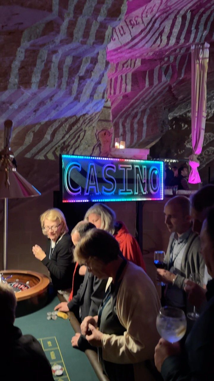 People gathered around a casino table near a glowing neon casino sign.