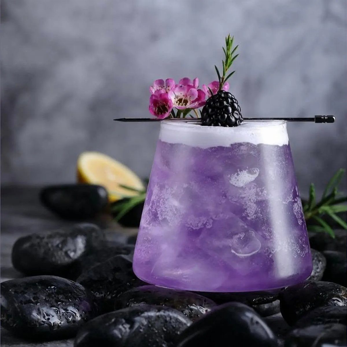 A purple cocktail with blackberry and floral garnish resting on black stones.