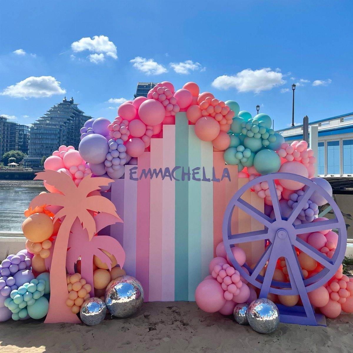 A pastel festival-themed backdrop with balloon arches, palm trees, and a purple Ferris wheel.