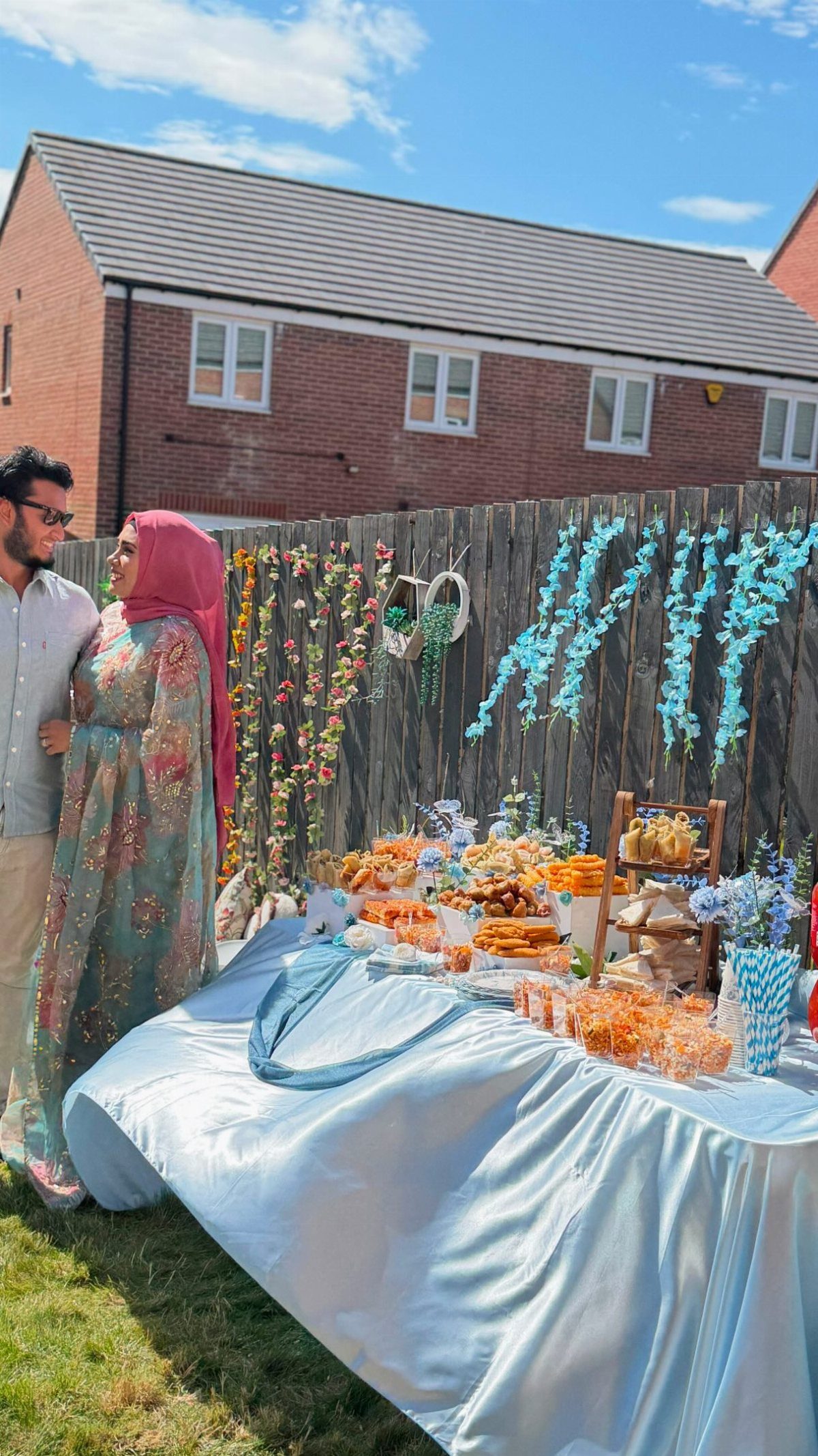 A man and woman standing beside a table of pastries and blue floral decorations outdoors.