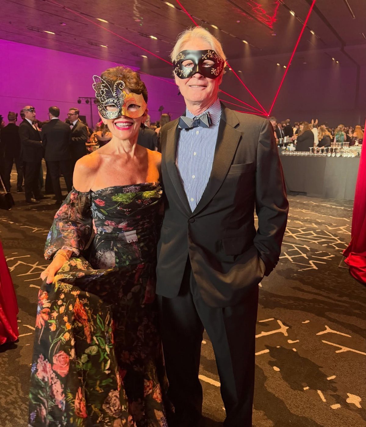 A man and woman wearing masquerade masks posing together in a large hall.