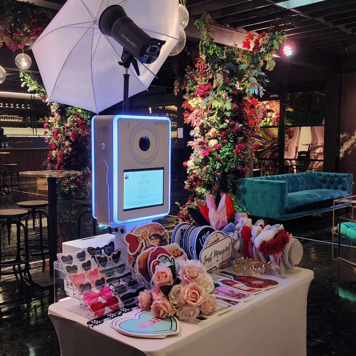A white photo booth machine with props and a photography umbrella in a decorated event space.