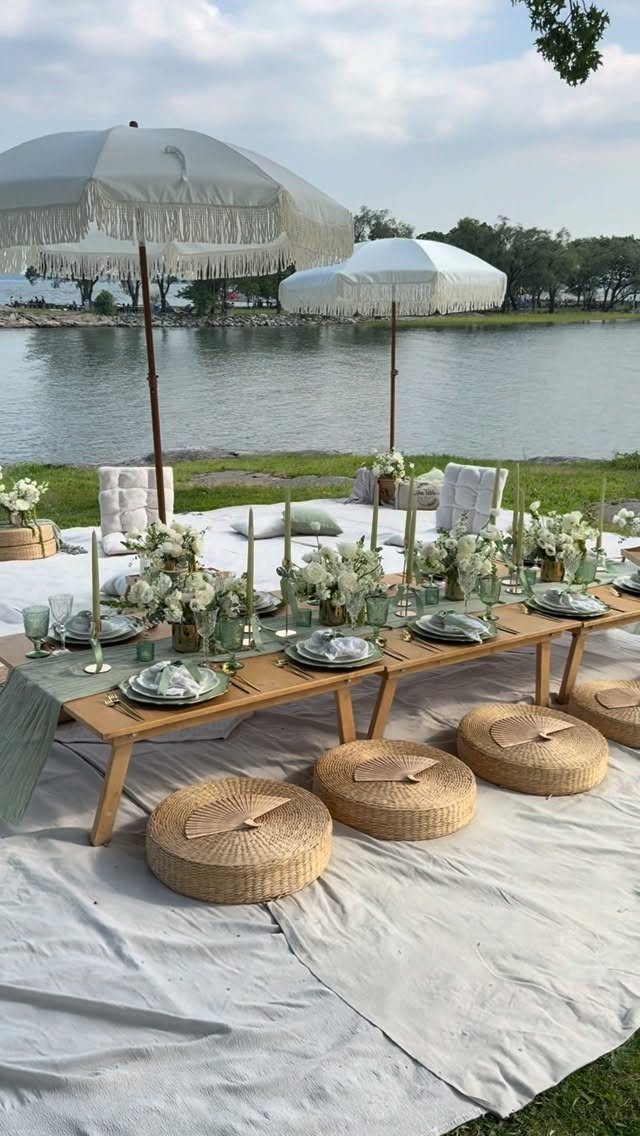 A luxury low-table picnic setup with white flowers and woven cushions by a lake.