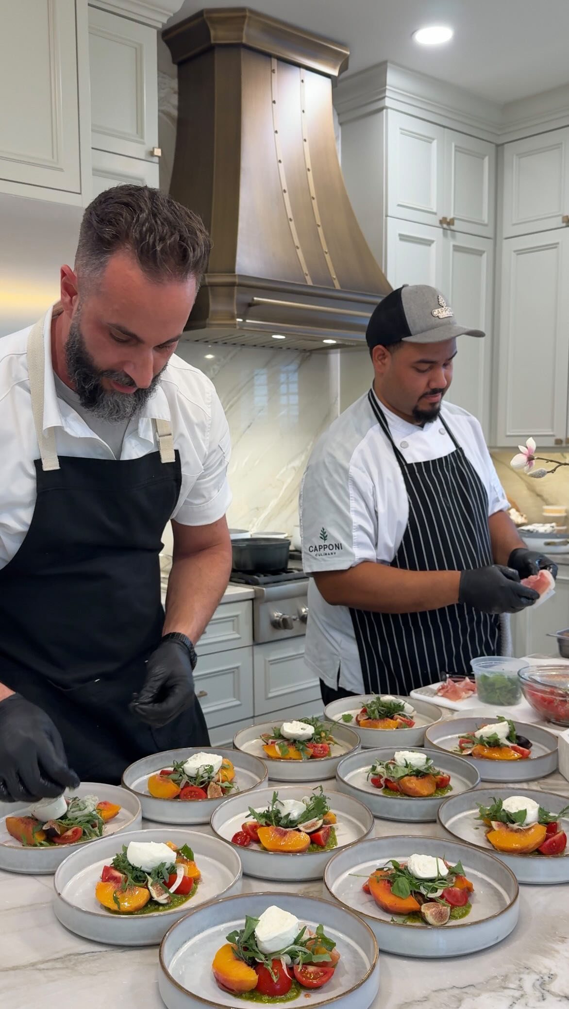 Two chefs in black aprons plating fresh peach and mozzarella appetizers in a modern kitchen.