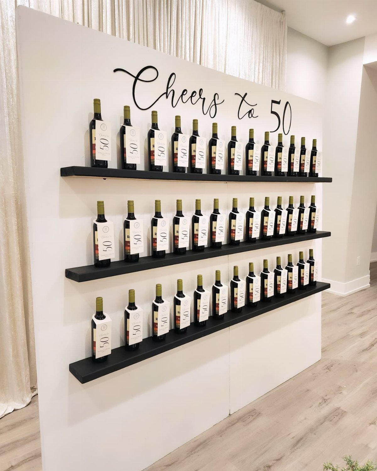 Wine bottles arranged on three black shelves against a white wall with Cheers to 50 text.