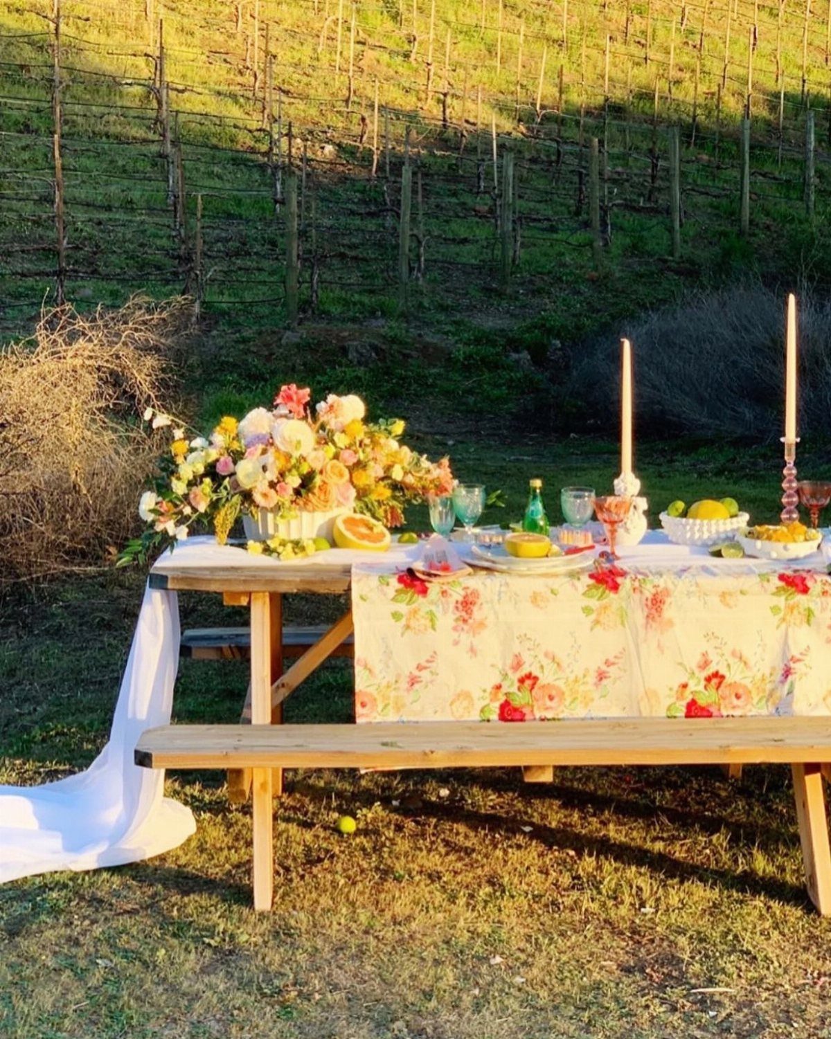 A decorated picnic table with floral tablecloth and flowers set against a vineyard background.