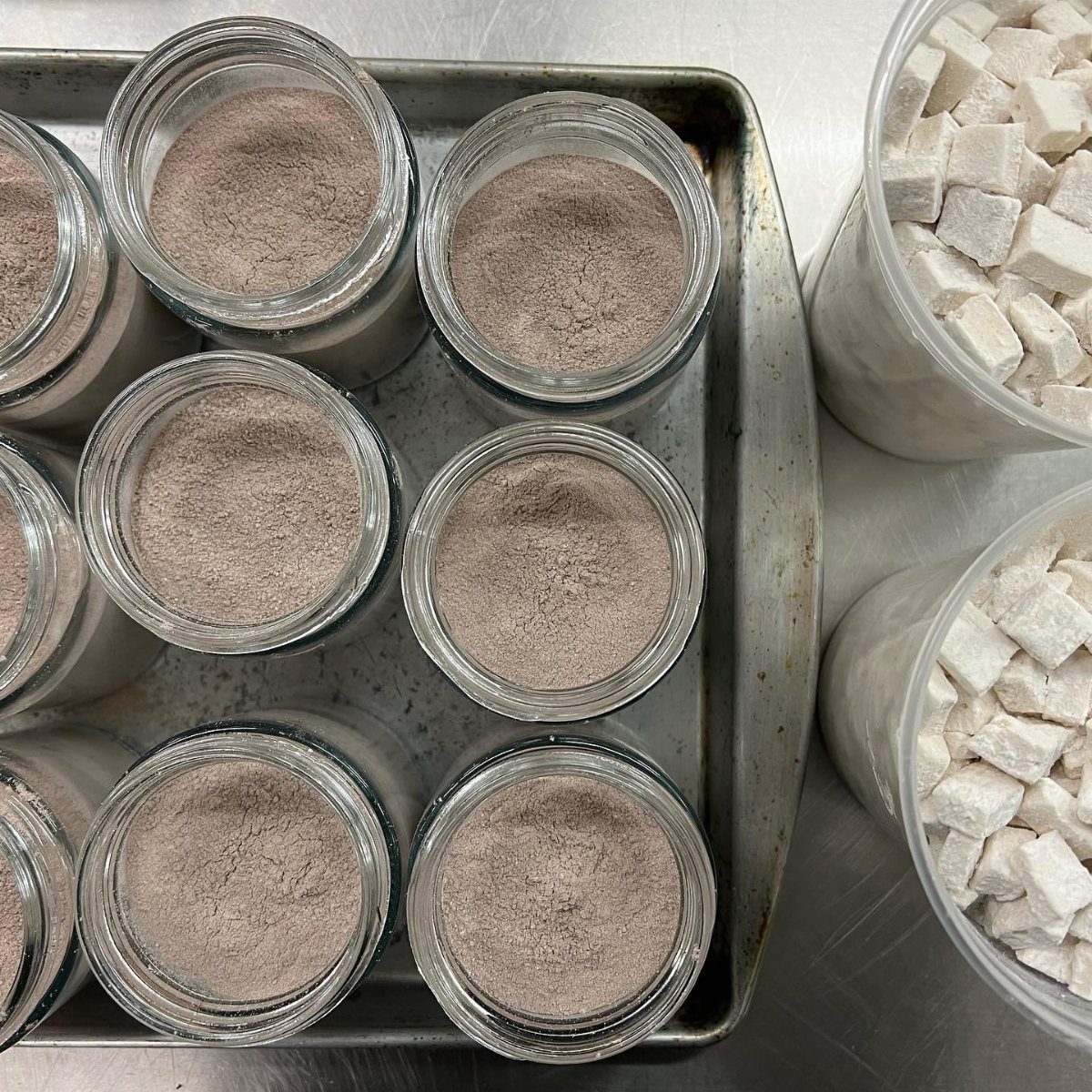 Glass jars filled with cocoa powder on a baking sheet next to containers of mini marshmallows.