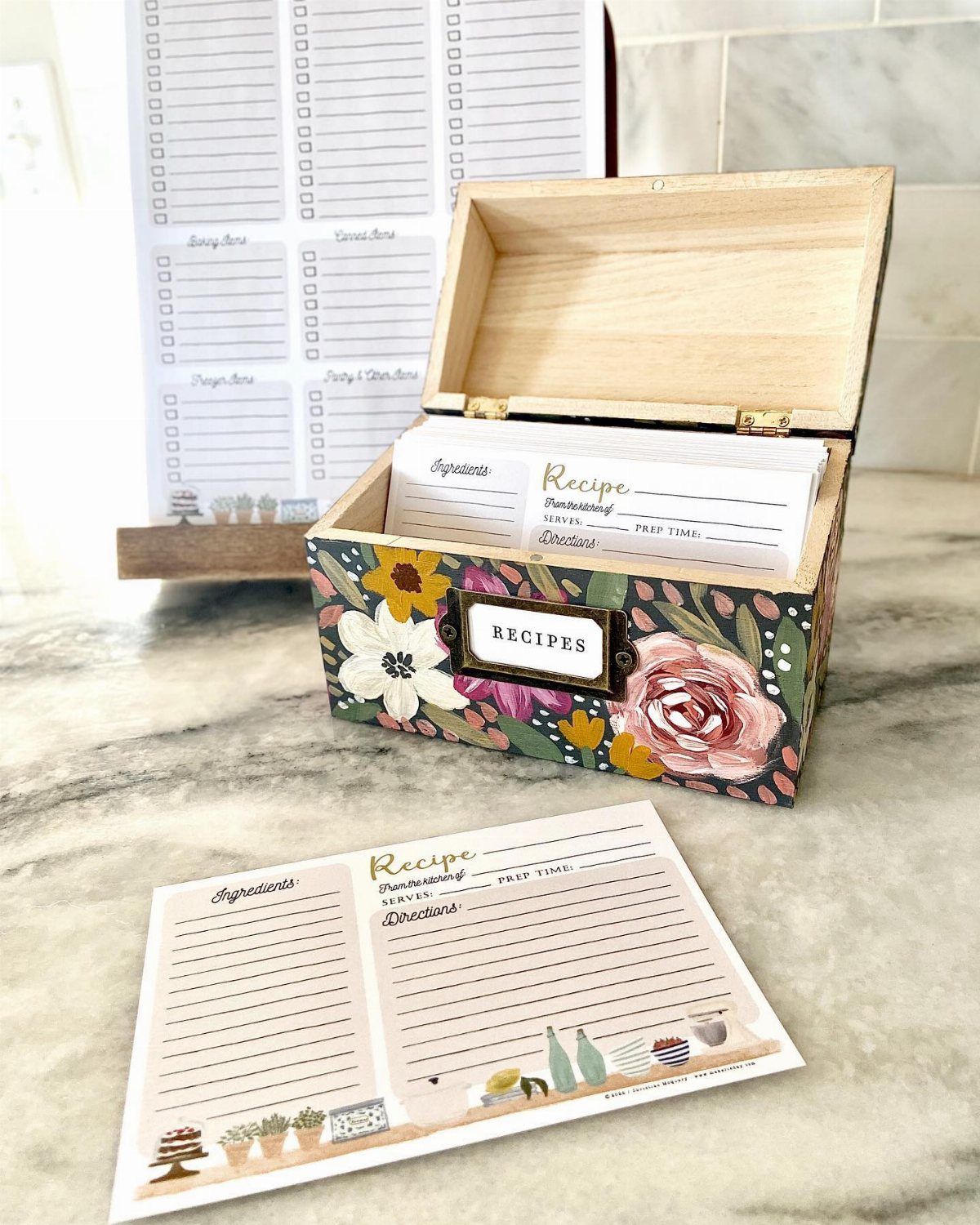 A floral wooden recipe box filled with cards and one recipe card lying on a marble surface.