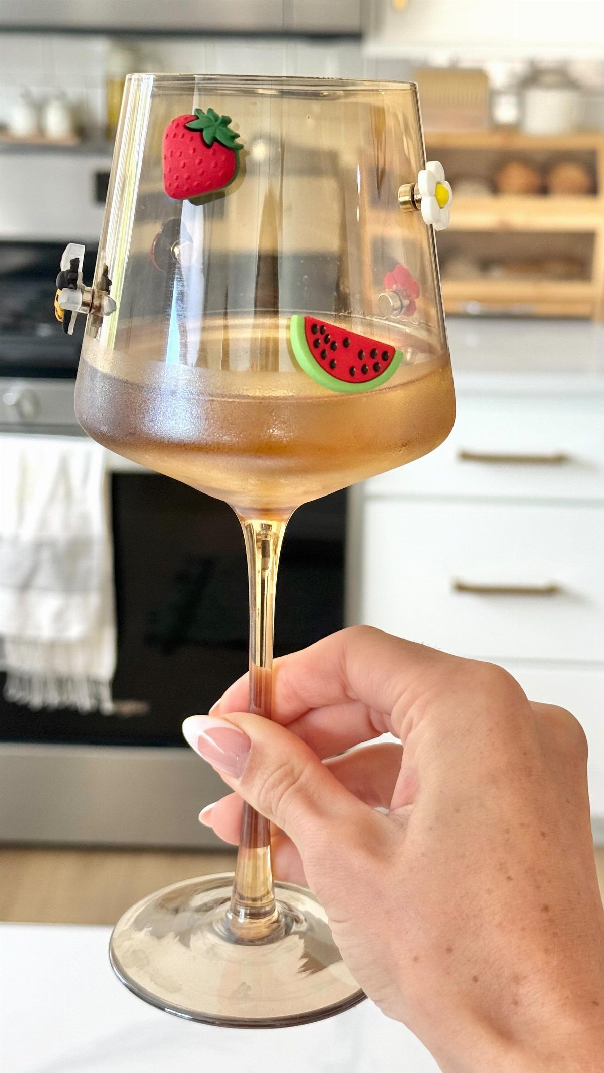 A hand holding an amber wine glass decorated with strawberry and watermelon charms.
