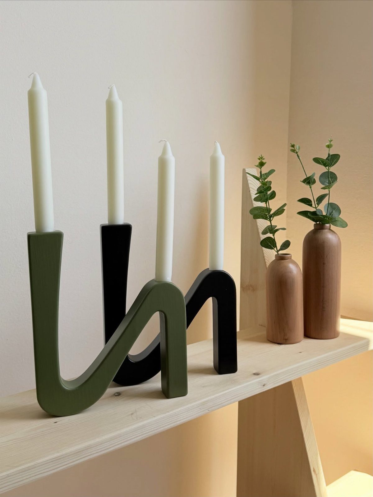 Modern wavy candle holders and small vases with eucalyptus on a wooden shelf.