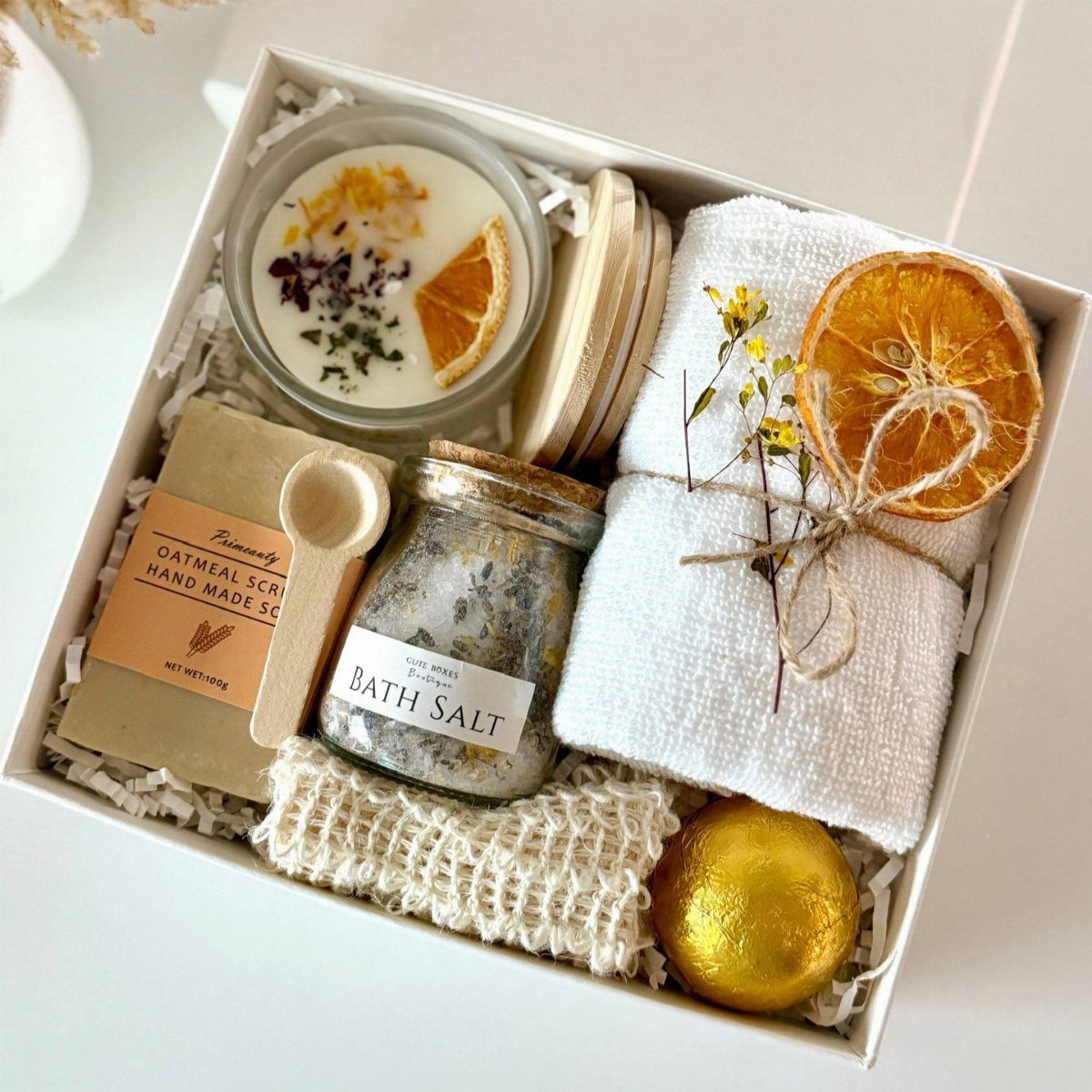 A white gift box containing a candle, bath salts, oatmeal scrub, and a rolled white towel.