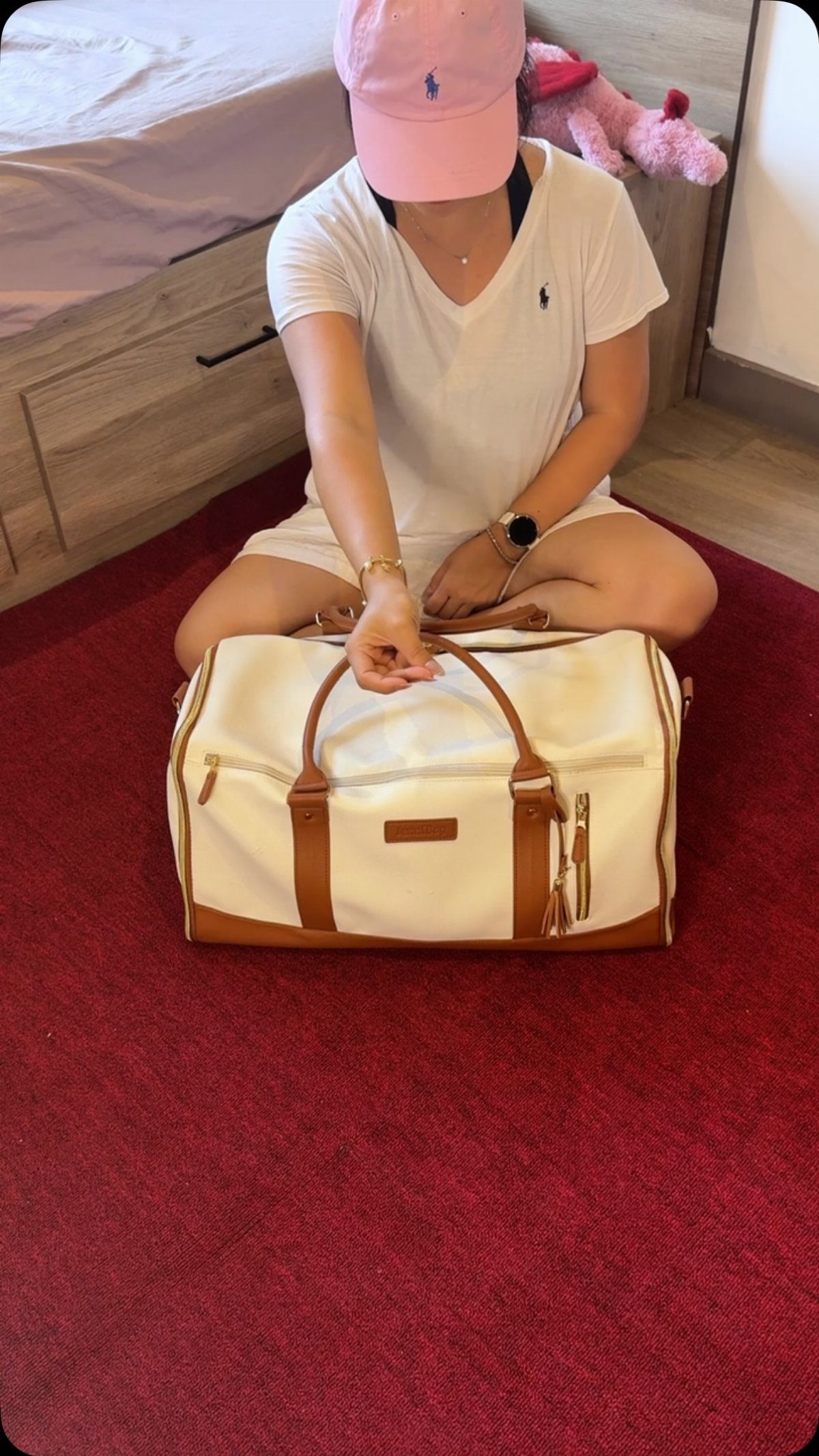 A person in a pink cap sits on a red rug next to a white duffel bag.