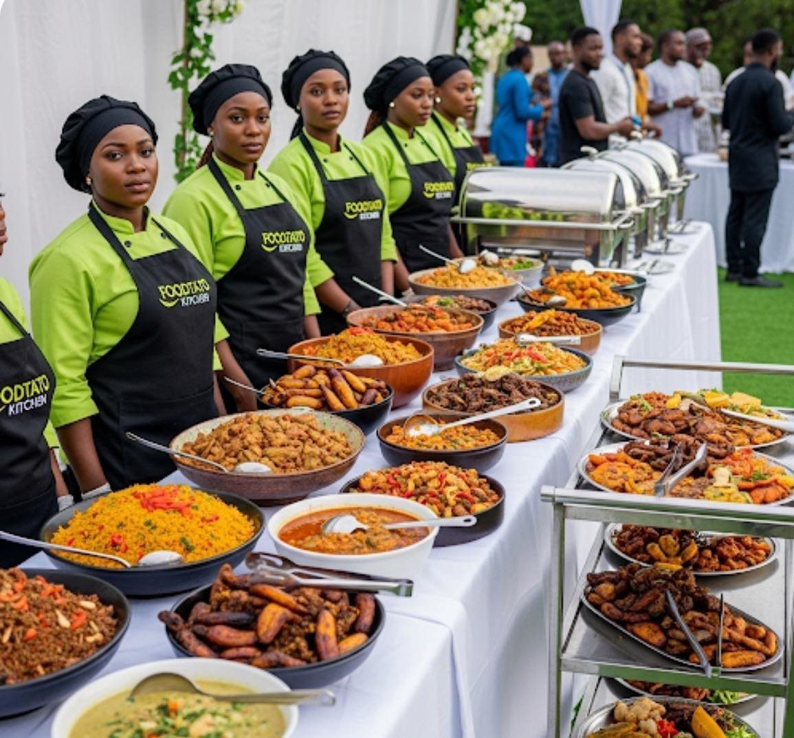 Caterers in green shirts and black aprons stand behind a long table filled with diverse buffet food.