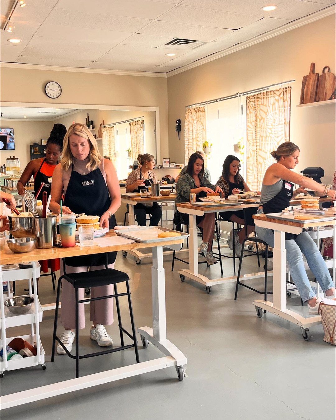 Women wearing aprons prepare food together at long white tables in a bright kitchen studio.