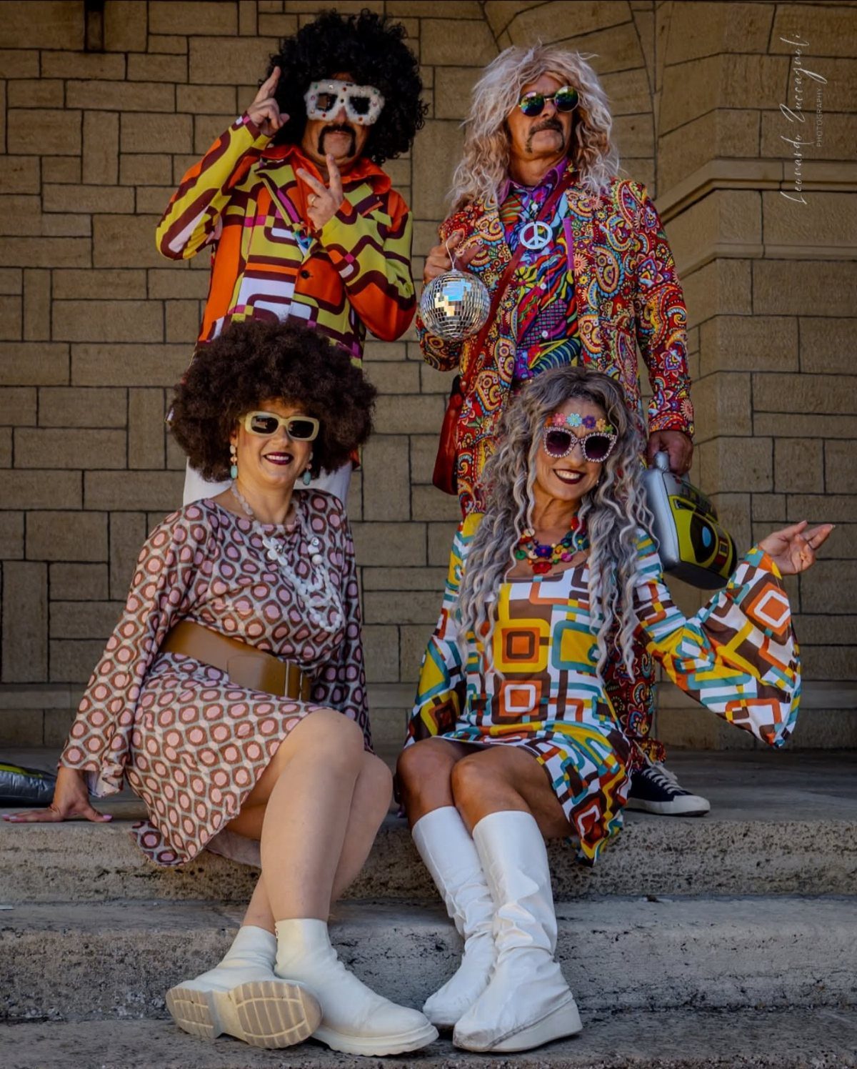 Four people in colorful 1970s disco attire posing on stone steps with a disco ball.
