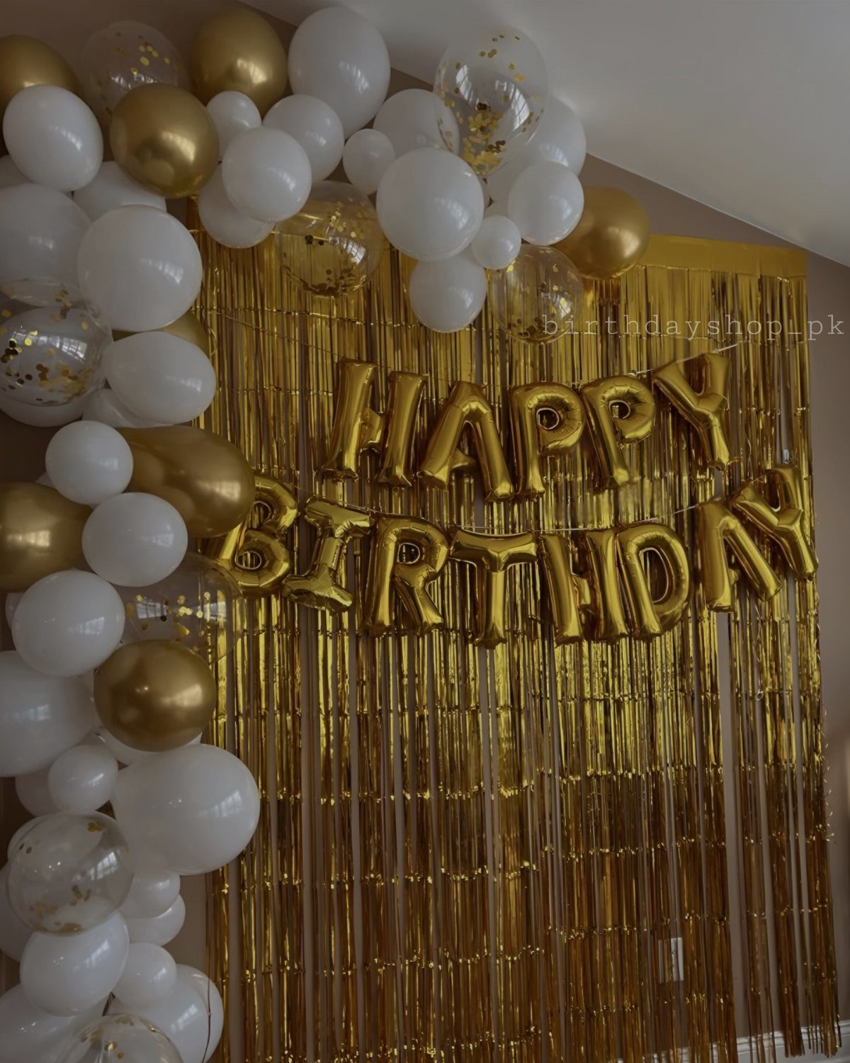 Gold and white balloons decorating a gold fringe curtain with Happy Birthday foil balloons.
