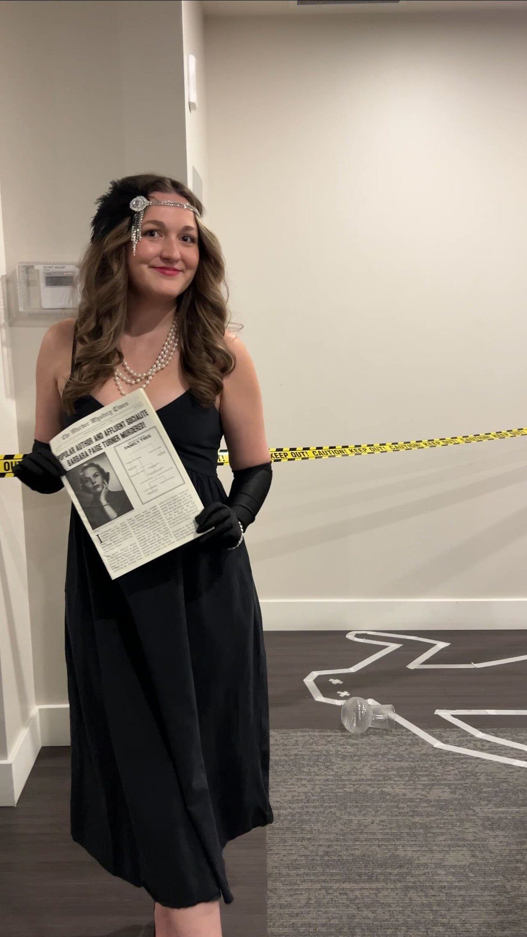 Woman in black evening attire holding a newspaper clipping while standing near yellow caution tape.