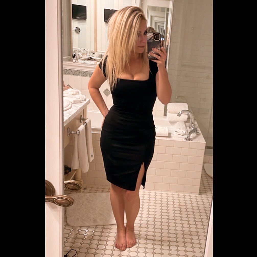 Blonde woman in a fitted black midi dress posing for a mirror selfie in a bathroom.