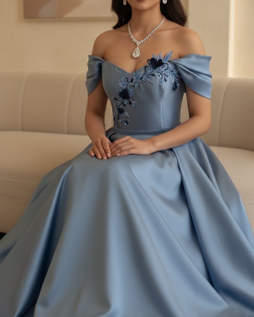 Woman in a light blue off-the-shoulder satin gown with floral embroidery sitting on a sofa.